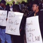 photo of male and female Black farmers at a protest, holding signs that read "Discrimination is wrong," "Don't take our land," and "Equal justice is the law"