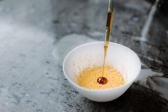 An artsy food shot shows a white bowl on a gray counter. A spatter of orange coats the bottom of the bowl while a device drips a syrupy dot on top. The orange is a fungus that gave this rice custard a fruity taste.