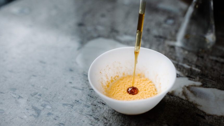 An artsy food shot shows a white bowl on a gray counter. A spatter of orange coats the bottom of the bowl while a device drips a syrupy dot on top. The orange is a fungus that gave this rice custard a fruity taste.