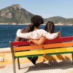 Stock photo, rear view of three multiethnic friends seated in an embrace on a colorful bench while looking out at the sea and mountains in the distance on a sunny day