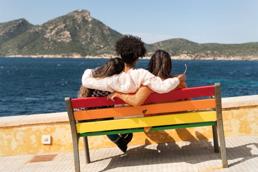 Stock photo, rear view of three multiethnic friends seated in an embrace on a colorful bench while looking out at the sea and mountains in the distance on a sunny day