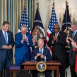 President Biden sits at a desk surrounded by other politicians and signs a bill