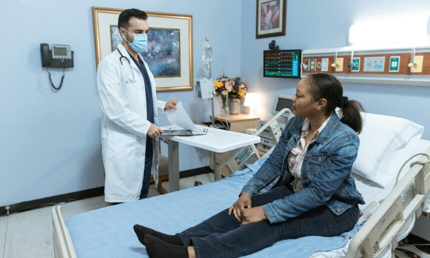 photo of a woman in street clothes sitting on a hospital bed talking to a doctor wearing a lab coat and mask and holding the patient's chart