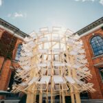 a pavilion in the courtyard of a museum made from dozens of white and timber folding deck chairs in a tall cylinder form