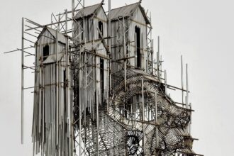 a wall sculpture made of numerous pieces of steel wire assembled in an abstract composition of house-like forms on scaffolding and the outline of a face