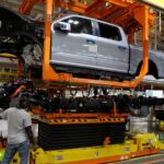 A man in a tee shirt and jeans raises a pickup truck on a piece of orange machinery in a factory