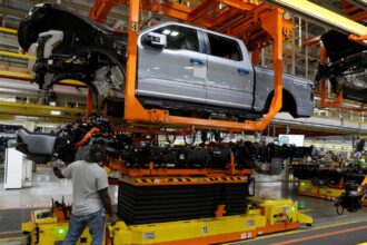 A man in a tee shirt and jeans raises a pickup truck on a piece of orange machinery in a factory