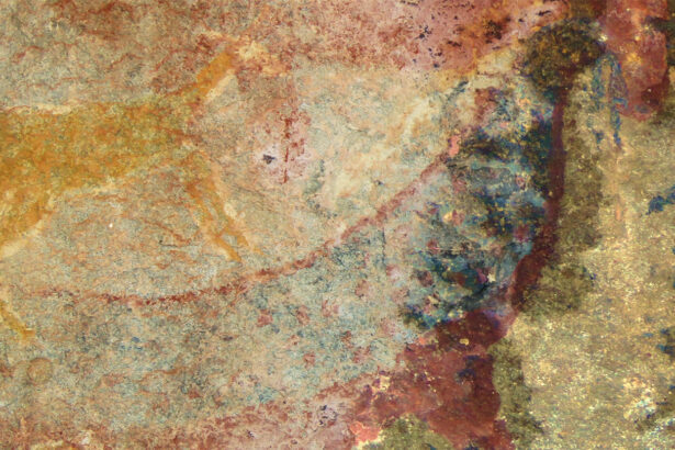 Faded paint on a cave wall appears to show two creatures, one in yellow top left and one in more reds and blues, that create an animal with an elongated body and what appears to be tusks, in blue, near its head region.