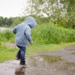 A kid wearing a gray hoodie, jeans, and sneakers splashes through a mud puddle outside on a gray day.