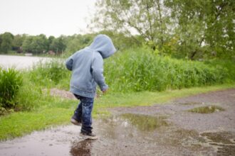 A kid wearing a gray hoodie, jeans, and sneakers splashes through a mud puddle outside on a gray day.