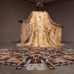 a large figure with a gold-yellow gown stands with outstretched arms. a circular display is on the floor in front of it