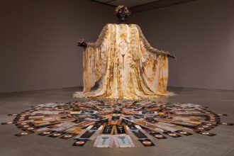 a large figure with a gold-yellow gown stands with outstretched arms. a circular display is on the floor in front of it