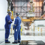 photo of two men in blue jumpsuits and hard hats talking inside the cavernous reactor hall of a nuclear power plant; there are caution signs and a radioactive symbol on the wall next to them