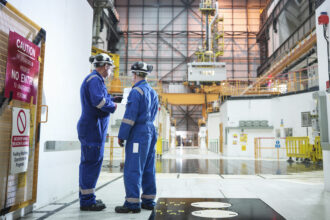 photo of two men in blue jumpsuits and hard hats talking inside the cavernous reactor hall of a nuclear power plant; there are caution signs and a radioactive symbol on the wall next to them