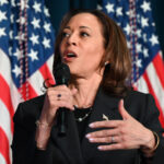 Photo of Kamala Harris speaking into a microphone with American flags in the background