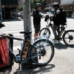 A person stands on a city street wearing a helmet and talking to another person, also helmeted, on a bike, with another bike with a red bag with the letters GH on it in the foreground