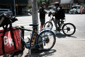 A person stands on a city street wearing a helmet and talking to another person, also helmeted, on a bike, with another bike with a red bag with the letters GH on it in the foreground