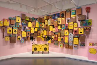 a collection of speakers painted with yellow and brown faces against a pink wall. a gramophone and boombox painted similarly stand in the front