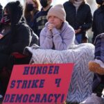 A coalition of conservative, progressive, and independent students, some over eight days into a hunger strike, congregate in front of the White House to urge President Biden to prioritize voting rights at Lafayette Park in December 2021 in Washington, DC. Centered is a young person holding a sign that reads Hunger Strike 4 Democracy!