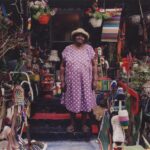 a Black woman in a pink and white polka dot dress and hat standing on the front step of her home with countless found objects sculptures surrounding her