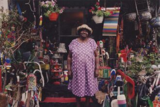 a Black woman in a pink and white polka dot dress and hat standing on the front step of her home with countless found objects sculptures surrounding her