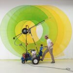 artist Thomas Trum moves a wheeled paint sprayer machine in front of a wall with green and yellow shapes being painted