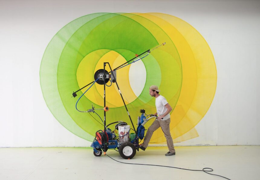 artist Thomas Trum moves a wheeled paint sprayer machine in front of a wall with green and yellow shapes being painted