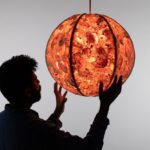 a male figure, silhouetted, reaches out to hold a glowing pendant lamp with a shade made out of sewn-together orange peels