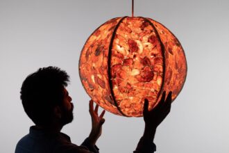 a male figure, silhouetted, reaches out to hold a glowing pendant lamp with a shade made out of sewn-together orange peels