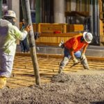 Two workers wearing hard hats spread concrete