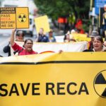 photo of protestors marching in a street, holding a yellow banner with radiation symbols and the words "save RECA"; other signs say "pass RECA before we die"