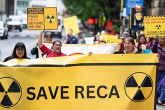 photo of protestors marching in a street, holding a yellow banner with radiation symbols and the words "save RECA"; other signs say "pass RECA before we die"