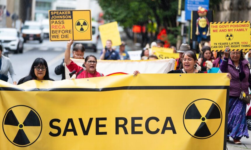 photo of protestors marching in a street, holding a yellow banner with radiation symbols and the words "save RECA"; other signs say "pass RECA before we die"