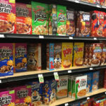 Grocery store shelves, filled with colorful cereals, like Raisin Bran, Apple Jacks, Pops, and Froot Loops.