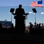 Republican presidential nominee  and former president Donald Trump, photographed from behind in silhouette, speaks from behind a glass barrier. In the distance beyond the silhouette of Trump, podium and stage, the audience and several American flags can be seen