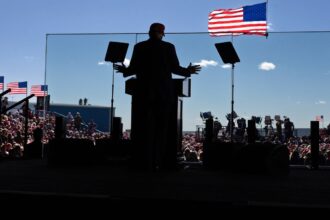 Republican presidential nominee  and former president Donald Trump, photographed from behind in silhouette, speaks from behind a glass barrier. In the distance beyond the silhouette of Trump, podium and stage, the audience and several American flags can be seen