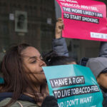 Two young people hold anti-tobacco signs at a a rally. The girl in front holds an teal-colored sign that reads, "I have a right to live tobacco-free." Another person holds a red sign that says, "The more tobacco ads I see, the more likely I am to start smoking."