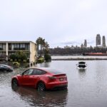 A red Tesla Model Y is partially submerged in the flooded parking lot of an apartment building.