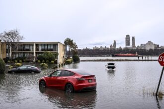 A red Tesla Model Y is partially submerged in the flooded parking lot of an apartment building.