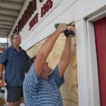 A man with a drill secures a wooden board over a store window in preparation for a hurricane to hit Florida