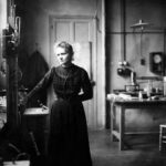 A black and white photograph showing Marie Curie standing in a science laboratory.