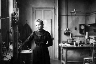 A black and white photograph showing Marie Curie standing in a science laboratory.