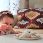 A little girl stands by a table reaching for a plate full of cookies.