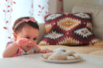 A little girl stands by a table reaching for a plate full of cookies.