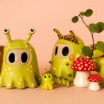 2 small ceramic ghosts posing a green snails and slug. one snail is significantly smaller and rests inbetween the two larger, implying it is a baby. Thet are surrounded by tiny red spotted mushrooms and a sprig with leaves