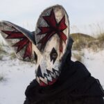 a portrait of a person wearing an ornate papier mache bat mask with red paint in the ears in a coastal setting