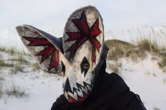 a portrait of a person wearing an ornate papier mache bat mask with red paint in the ears in a coastal setting