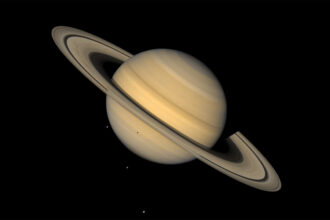 This image shows Saturn, ringed and in tones of tan, against an inky background. Three tiny light spots in the background and one dot against the lower half of the planet are four of Saturn