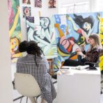 Two people sitting and conversing in front of bright, abstracted figurative paintings.