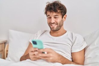 A happy guy sitting in bed reading on his phone.